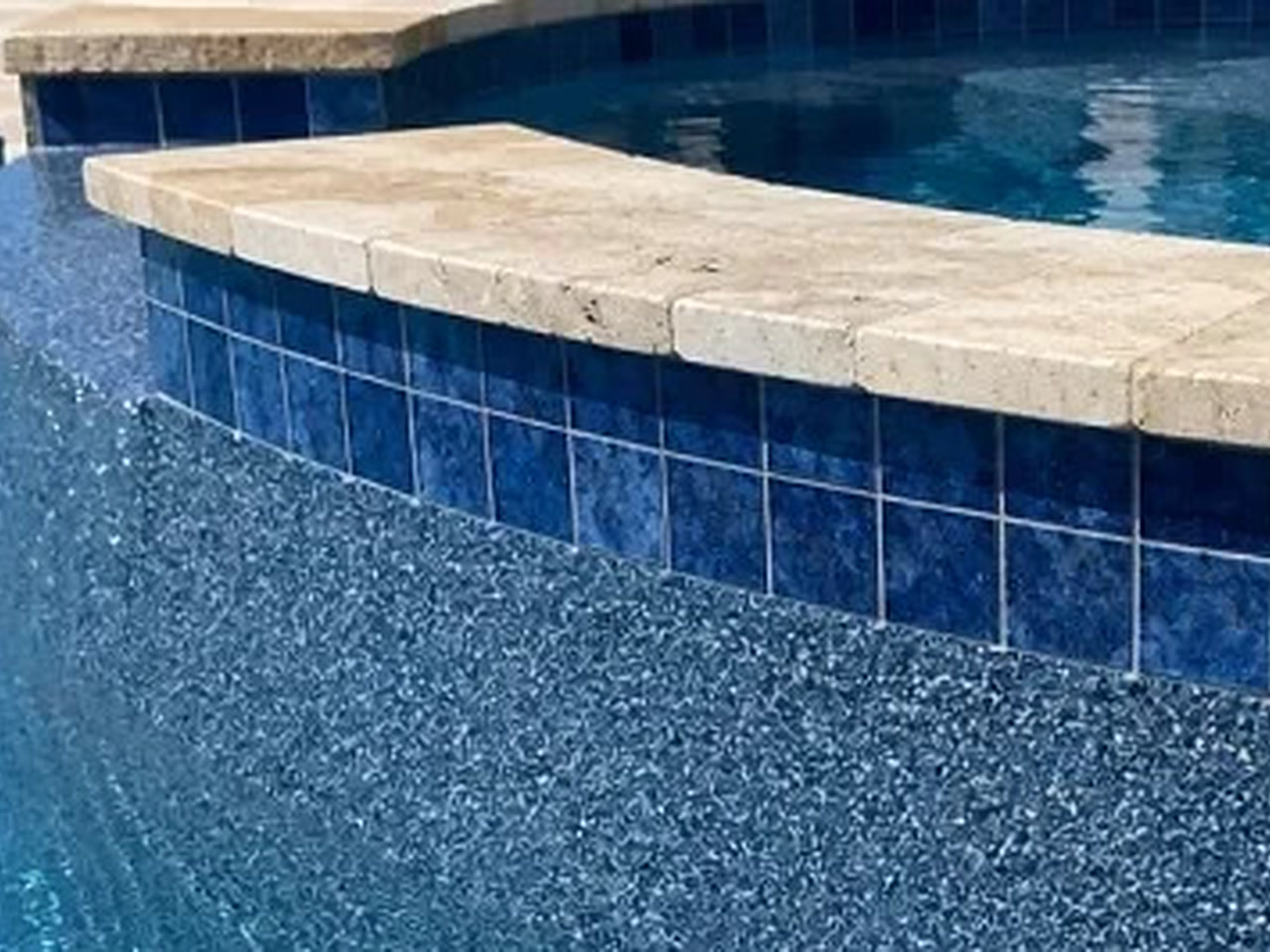 Blue pebble pool finish meeting a raised spa wall of deep blue tile capped with travertine coping on a West Lake property