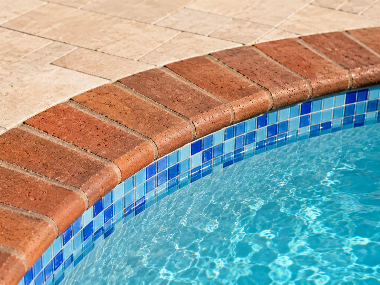 Red brick pool coping following a curved edge over blue mosaic waterline tile, beside a travertine deck at West Lake