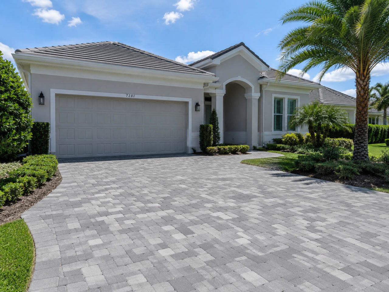 Gray tumbled paver driveway curving up to the two car garage of a Florida home framed by clipped hedges and palms
